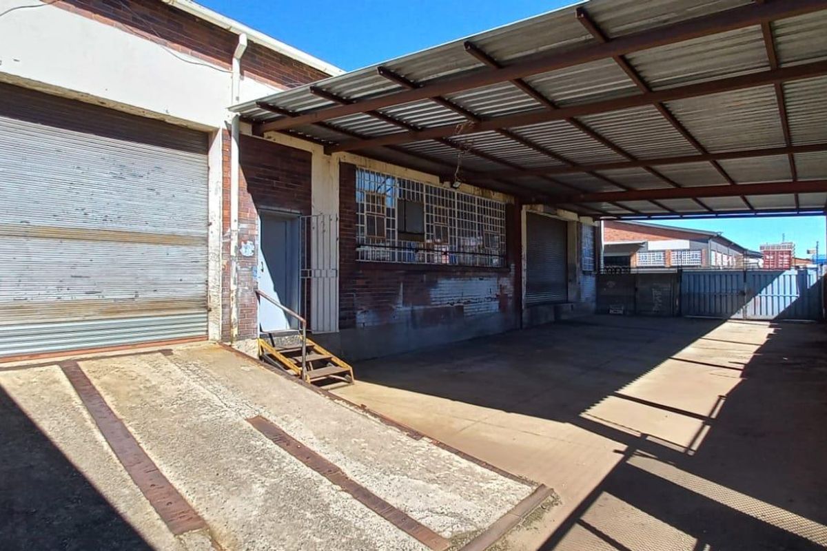 Industrial exterior view featuring a covered loading bay, concrete ramp, roller shutter doors, and brick facade with security bars.