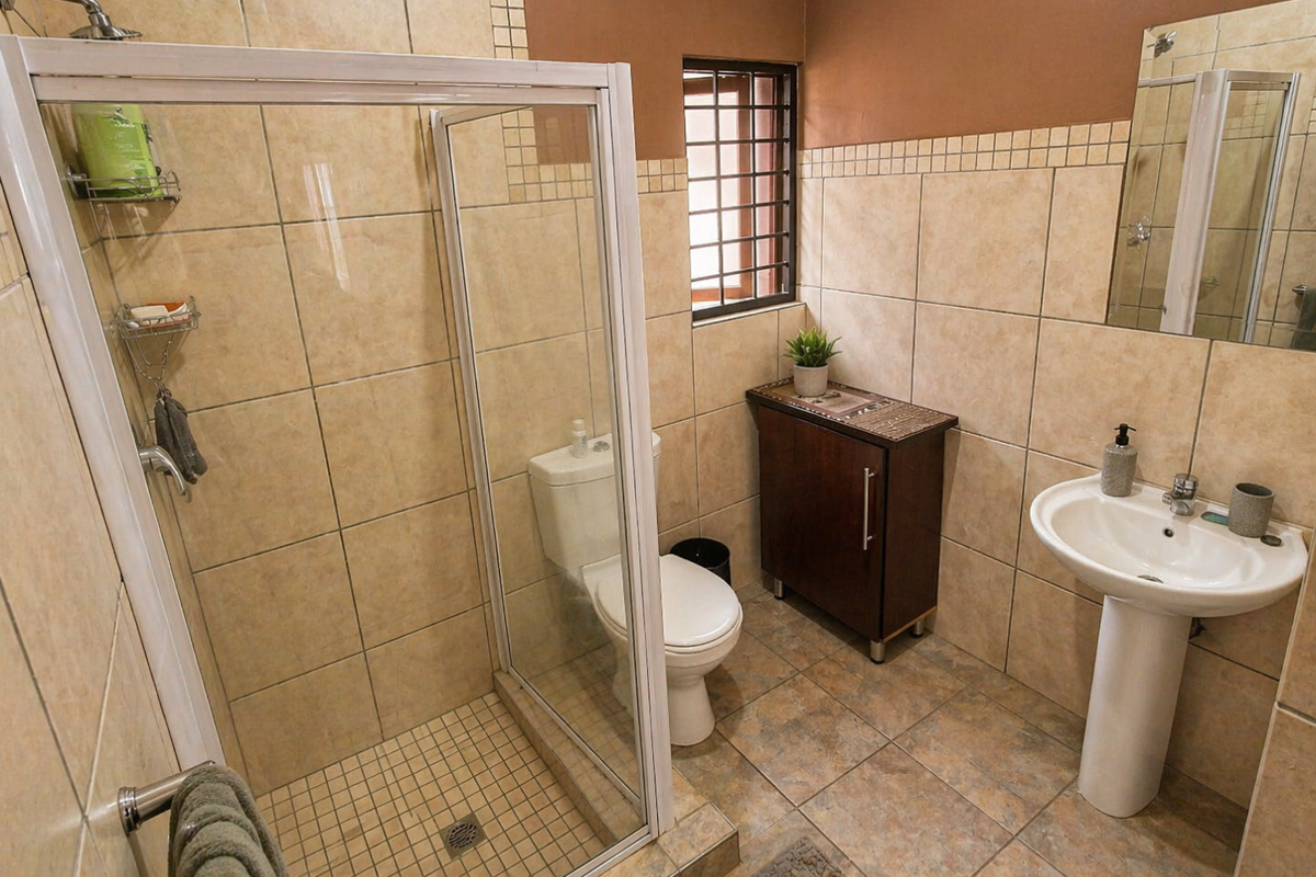 A functional bathroom featuring a glass-enclosed shower, pedestal basin, and toilet with neutral wall tiling and a security-barred window.