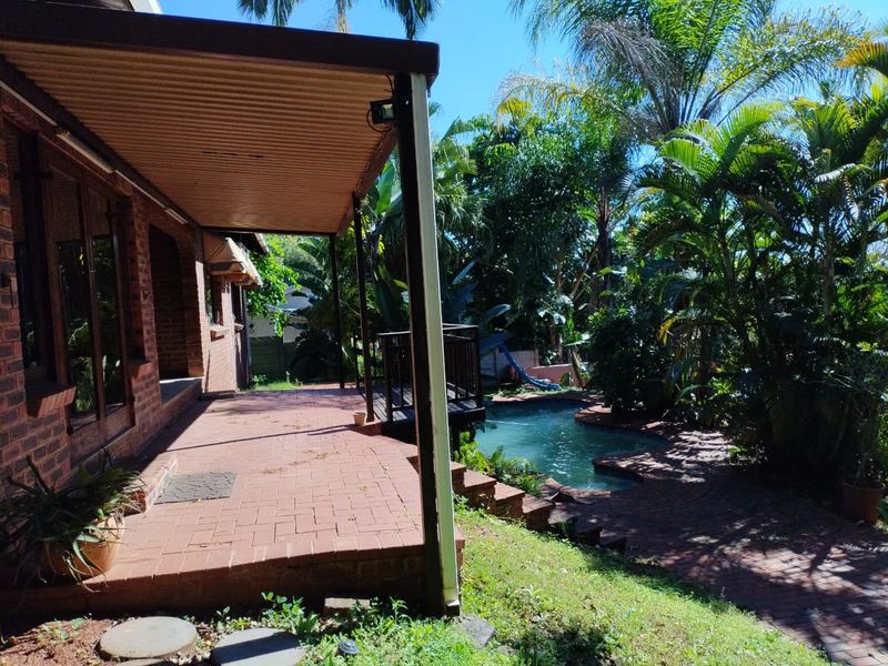 Outdoor entertainment area featuring a covered brick patio overlooking a swimming pool and lush garden surroundings.