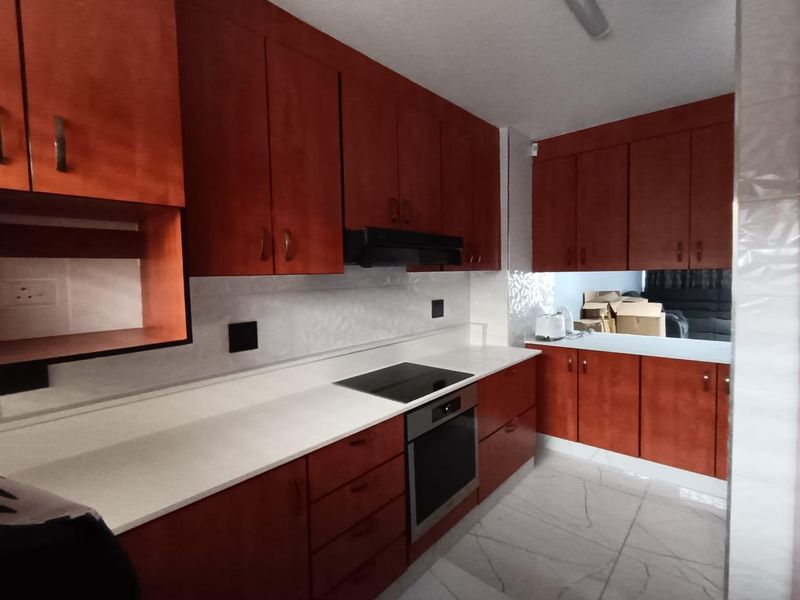 A functional kitchen featuring warm-toned wooden cabinetry, stone countertops, a built-in electric hob, oven, and modern tiled flooring.