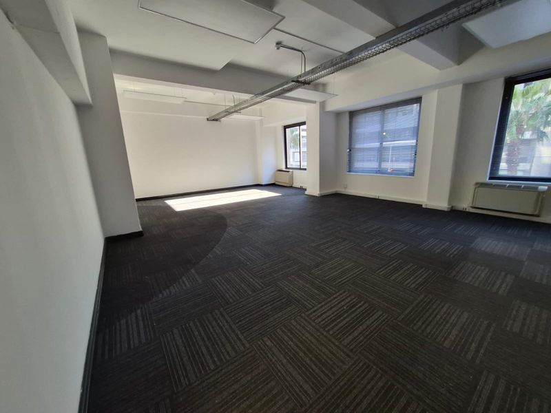 A spacious, vacant open-plan office area featuring dark carpet tiles, exposed industrial cable trays, and large windows with venetian blinds.
