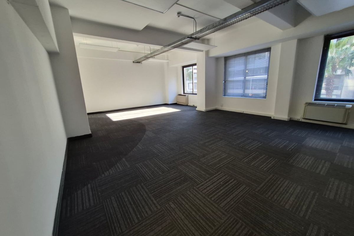 A spacious, vacant open-plan office area featuring dark carpet tiles, exposed industrial cable trays, and large windows with venetian blinds.