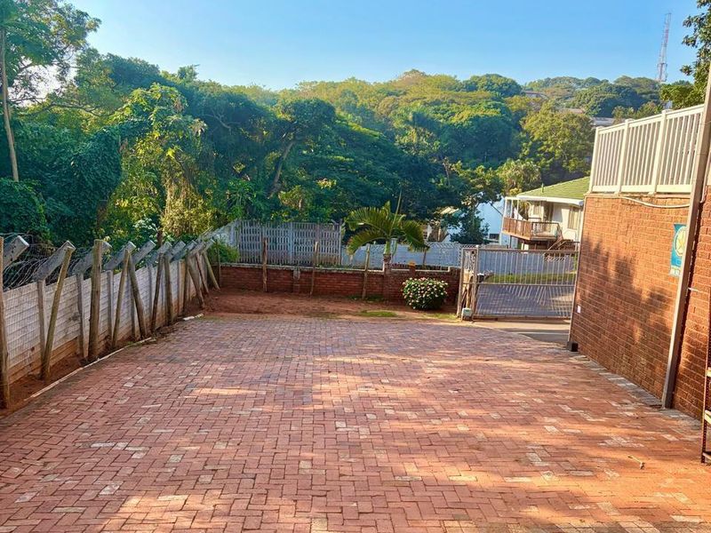 A paved driveway area featuring brick retaining walls, security fencing, and a metal gate, set against a backdrop of lush greenery.