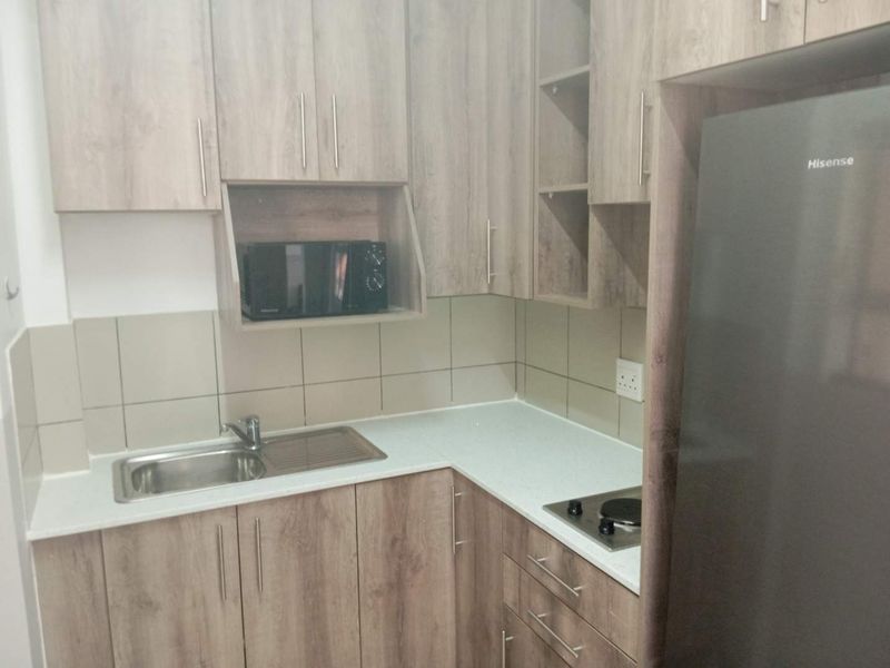 A compact, modern kitchen featuring light wood-grain cabinetry, stone countertops, a stainless steel sink, and a built-in hob with tiled backsplash.