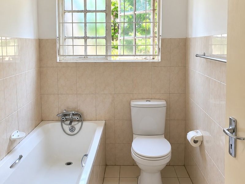 A functional bathroom featuring a standard bathtub, toilet, and neutral wall and floor tiling, complemented by a window with security bars.