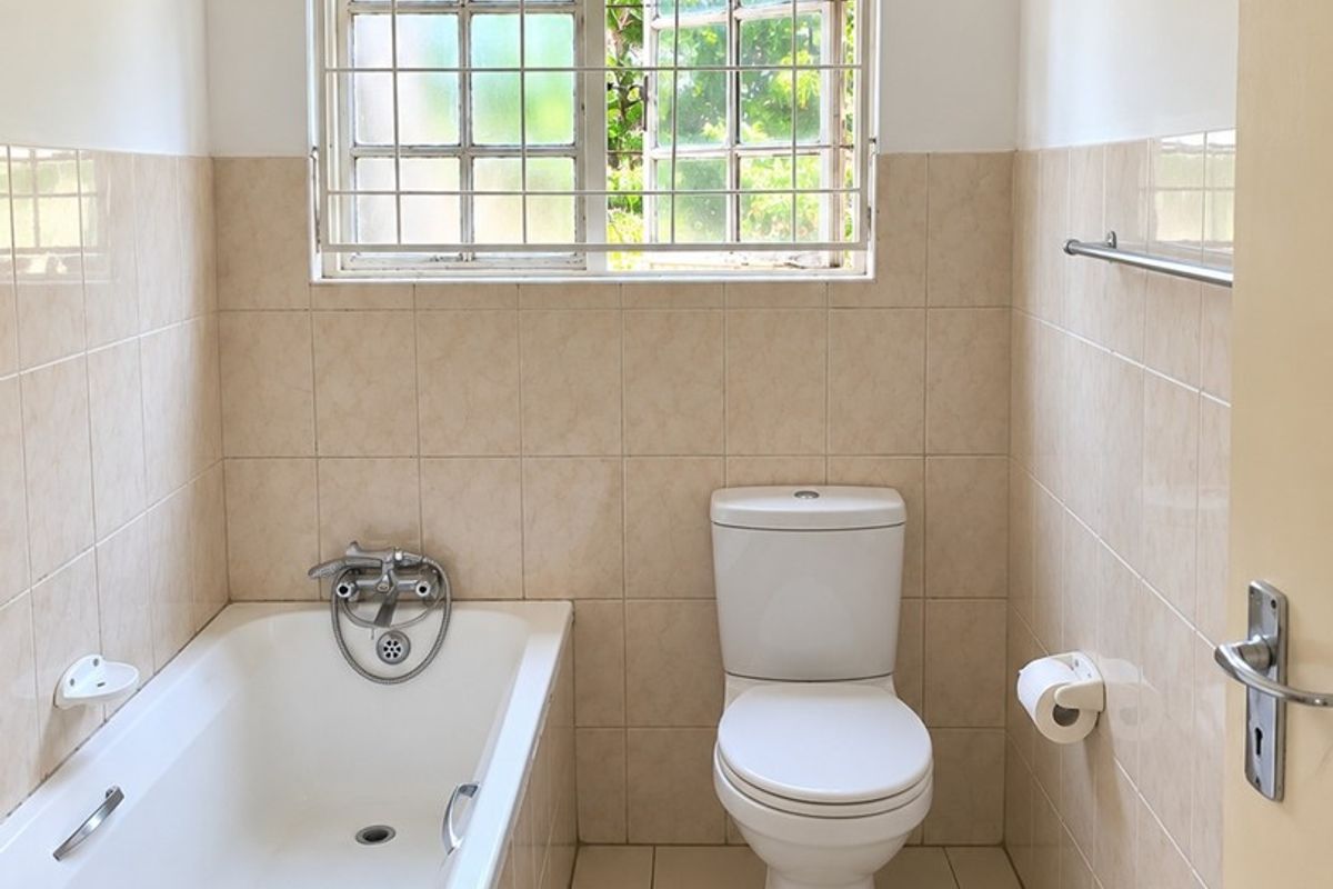 A functional bathroom featuring a standard bathtub, toilet, and neutral wall and floor tiling, complemented by a window with security bars.