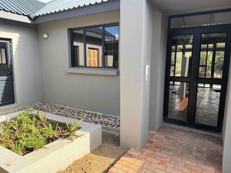 A modern exterior entrance featuring a brick paved walkway, a raised concrete planter, and dark aluminium framed glass doors and windows.
