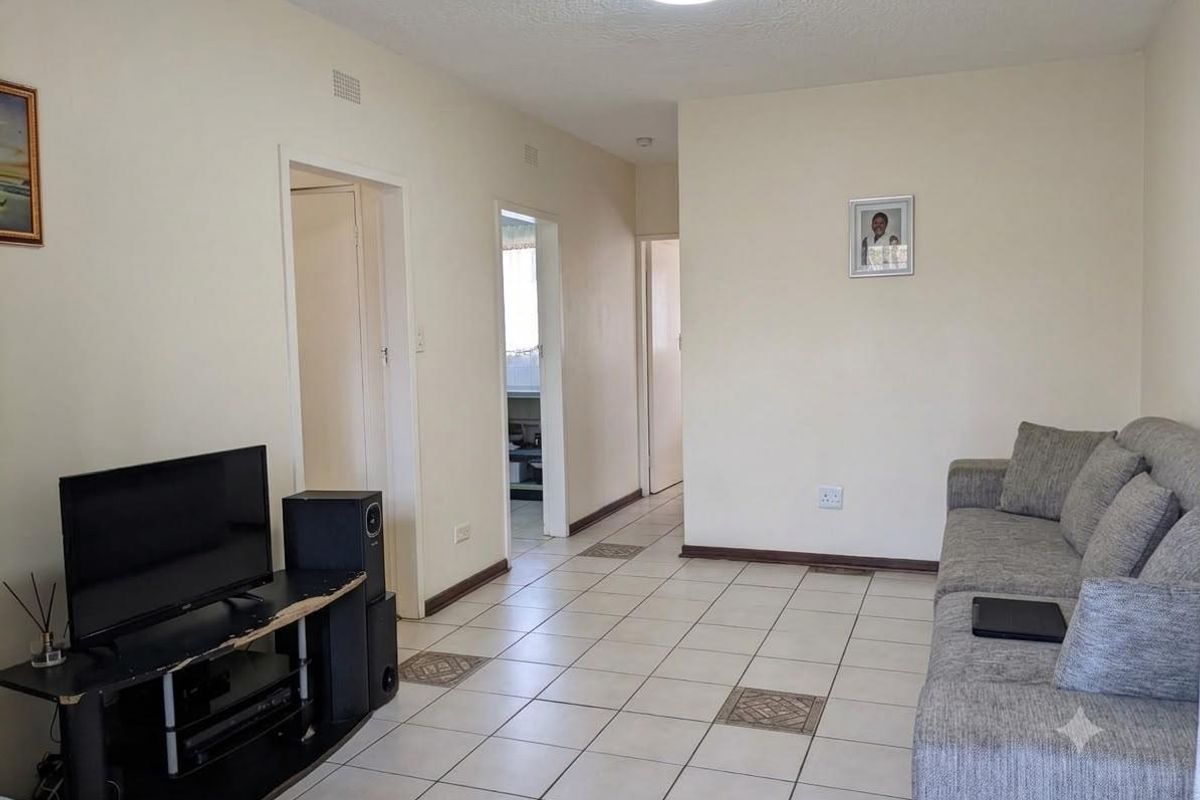 A functional living room featuring neutral ceramic floor tiles, standard ceiling lighting, and access to adjacent rooms via a central hallway.