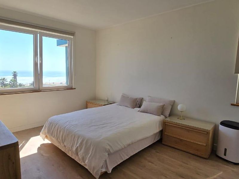 A bright bedroom featuring light-toned laminate flooring, roller blinds, and large windows offering a scenic coastal view.