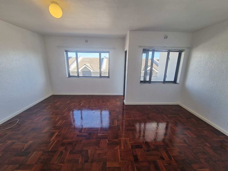 A bright, empty living room featuring polished parquet flooring, white walls, and large windows providing natural light.