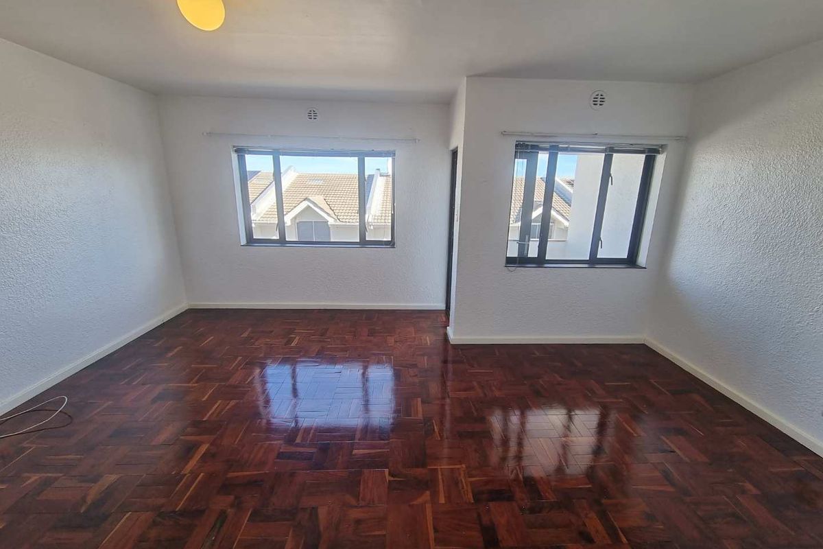 A bright, empty living room featuring polished parquet flooring, white walls, and large windows providing natural light.