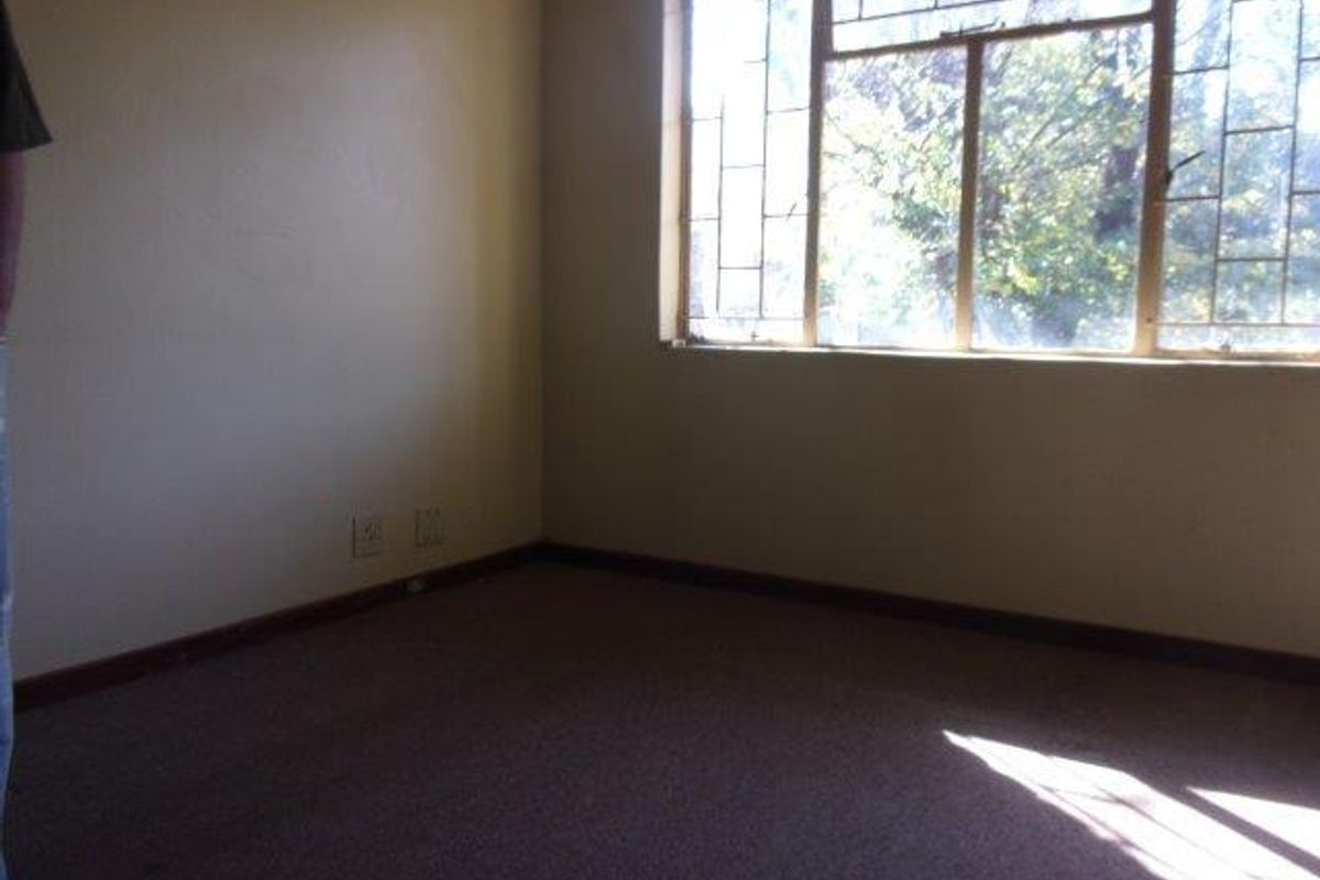 An empty bedroom featuring carpet flooring and a large window with security bars, offering natural light and a view of the exterior greenery.