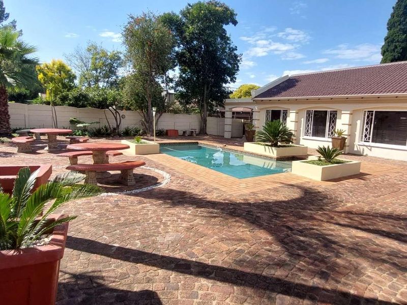 A well-maintained outdoor pool area featuring a paved patio, raised garden beds, and a swimming pool, set against the rear facade of the house.
