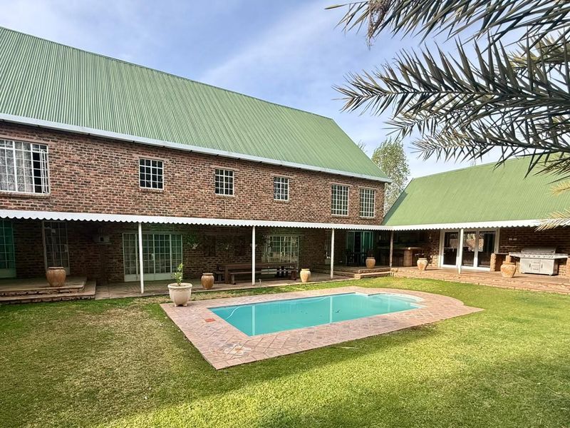 Spacious outdoor entertainment area featuring a swimming pool, paved surround, and a large brick dwelling with a covered patio and built-in braai.