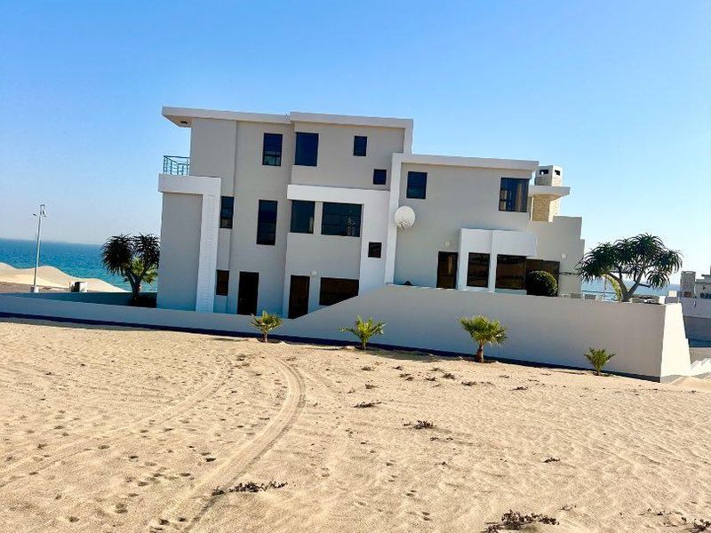 A modern multi-story coastal residence featuring clean architectural lines, a balcony, and a boundary wall, situated on a sandy beachfront plot.