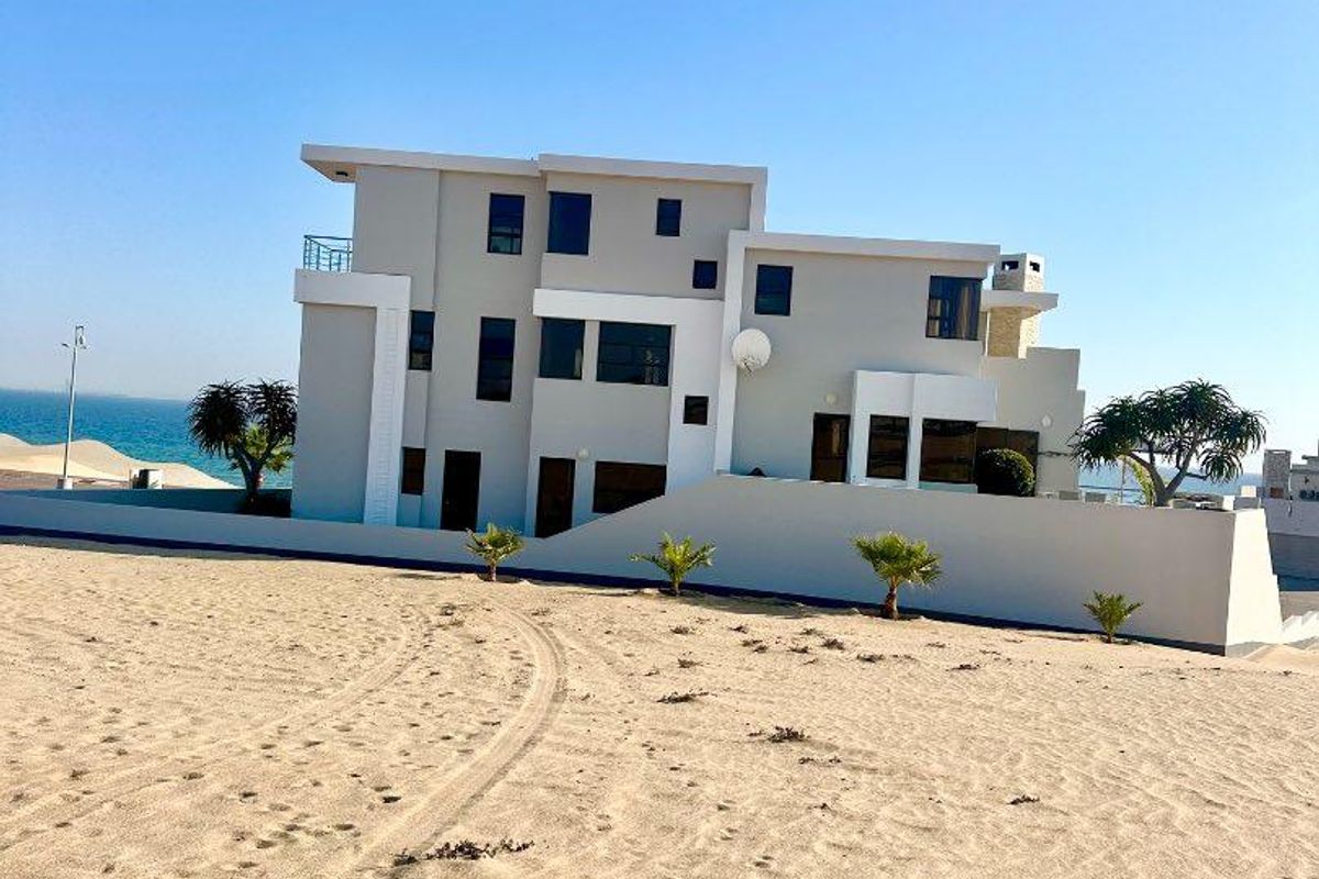 A modern multi-story coastal residence featuring clean architectural lines, a balcony, and a boundary wall, situated on a sandy beachfront plot.