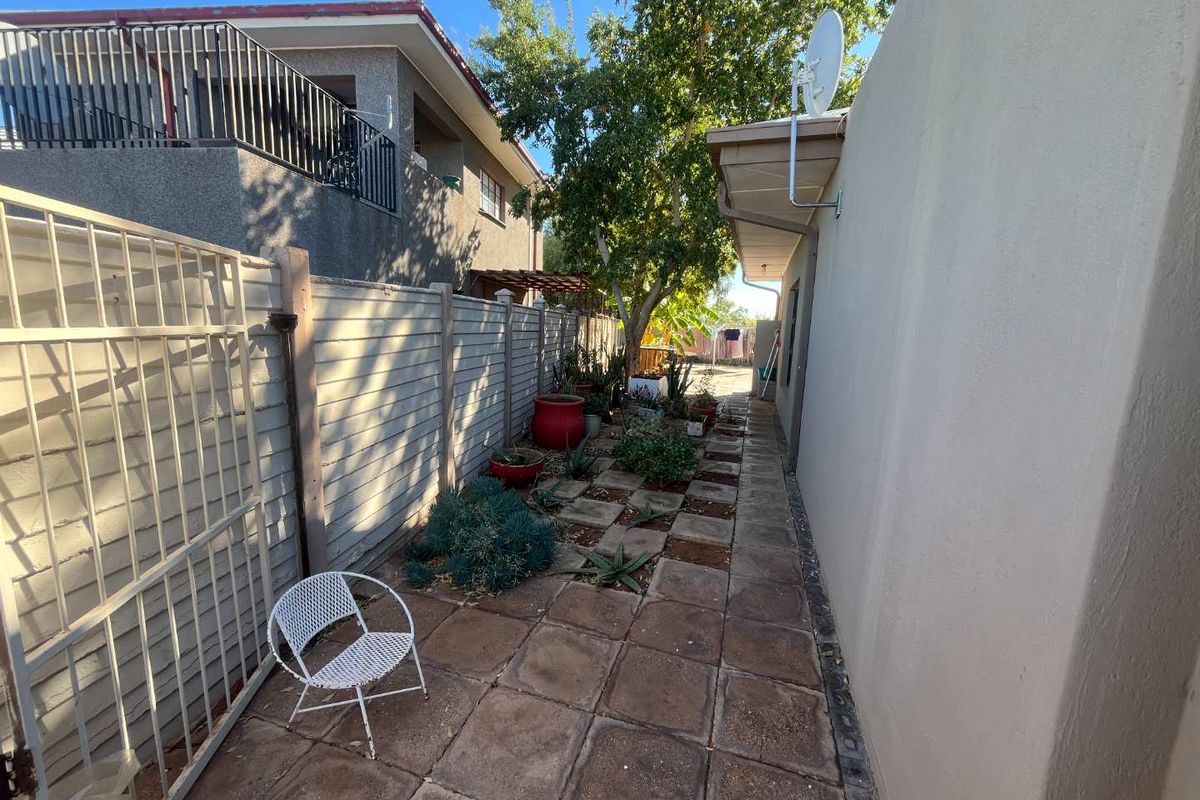 A narrow side garden area featuring paved walkways, established trees, and a secure metal gate along the side of the property.