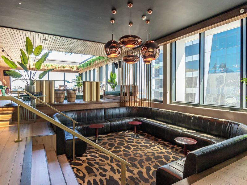 A sophisticated clubhouse lounge area featuring tiered timber flooring, integrated recessed lighting, large floor-to-ceiling windows, and modern metallic pendant light fixtures.