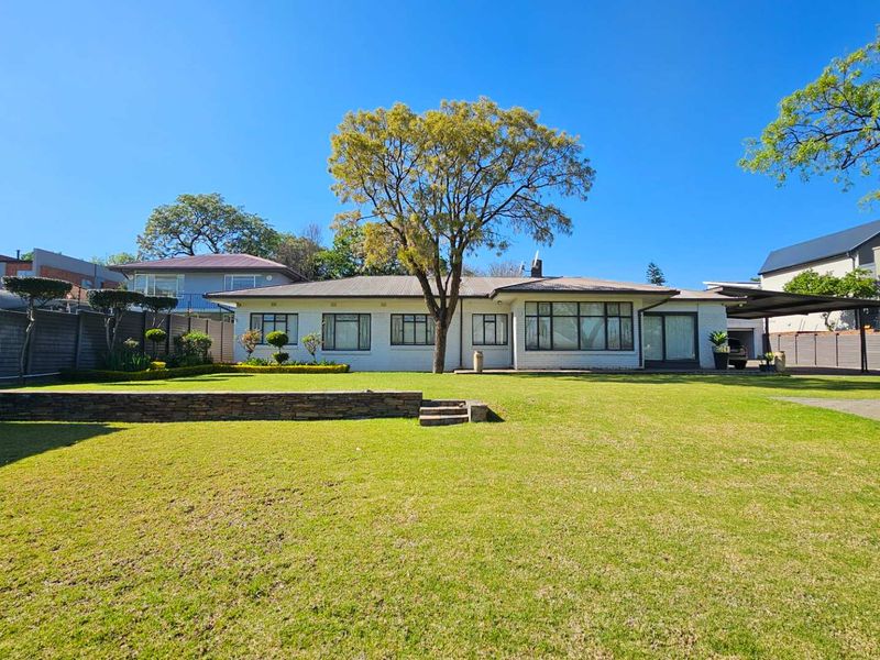 Spacious rear garden featuring a well-maintained lawn, stone retaining wall, and a paved driveway leading to a carport.