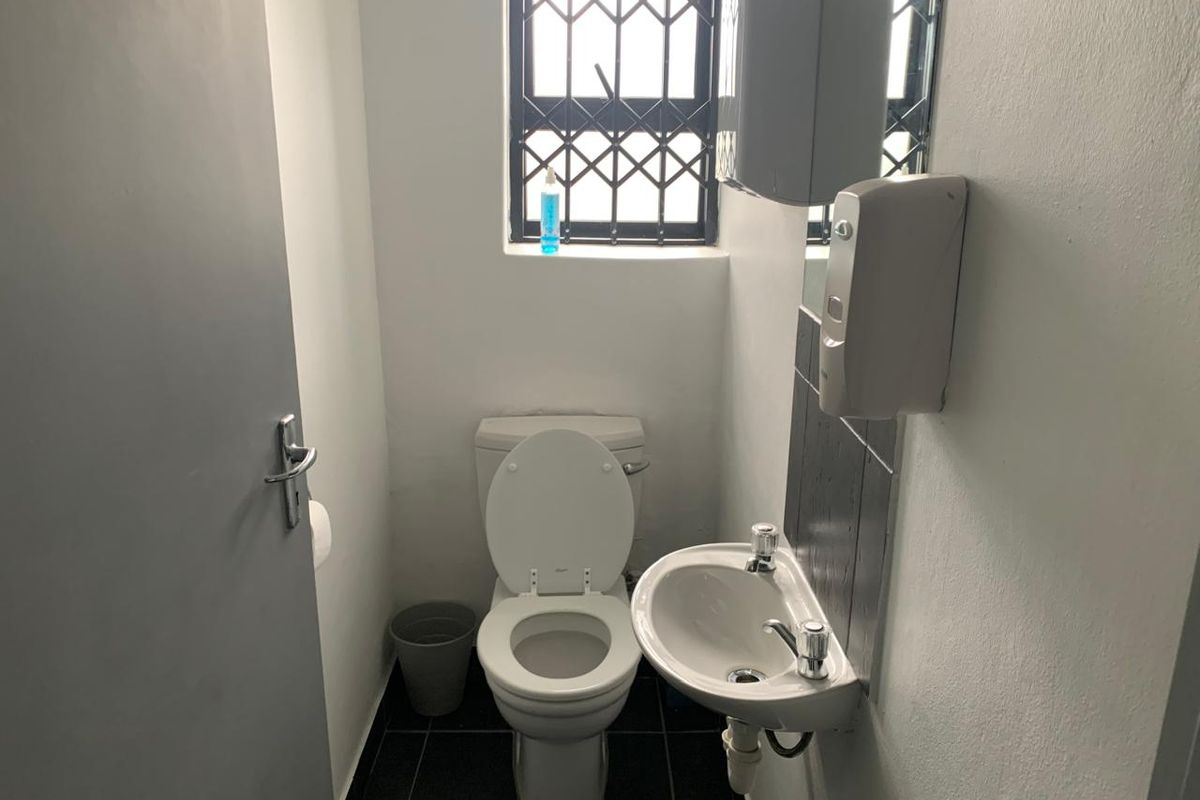 A compact industrial restroom featuring a wall-mounted basin, a standard toilet, and a security-grille window.