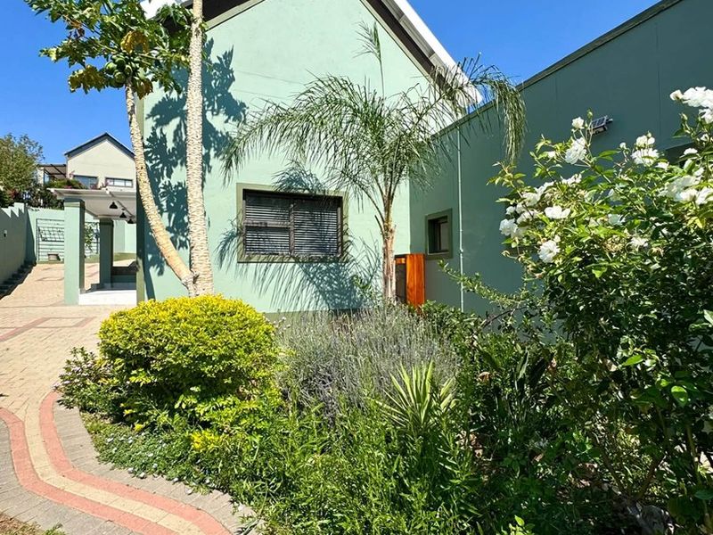 A well-maintained rear exterior view featuring a paved walkway, landscaped garden beds, and a light green facade with a gable roof.