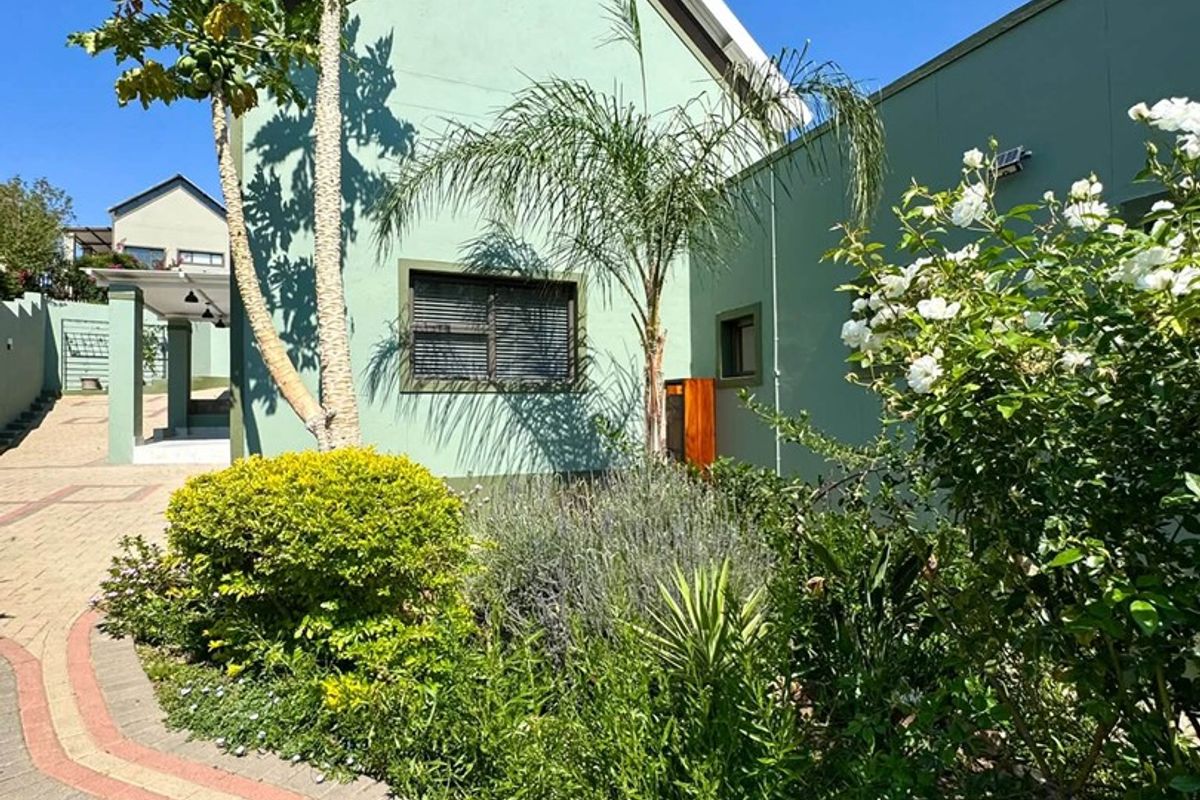 A well-maintained rear exterior view featuring a paved walkway, landscaped garden beds, and a light green facade with a gable roof.