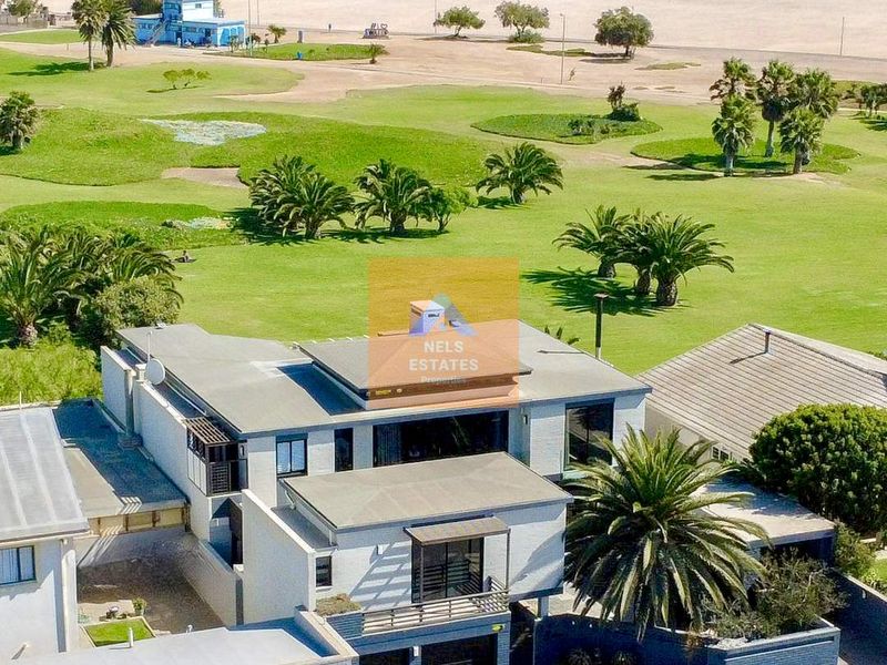 Aerial view of a modern multi-level coastal residence featuring a flat roof, balcony, and double garage, situated near the beach and golf course.