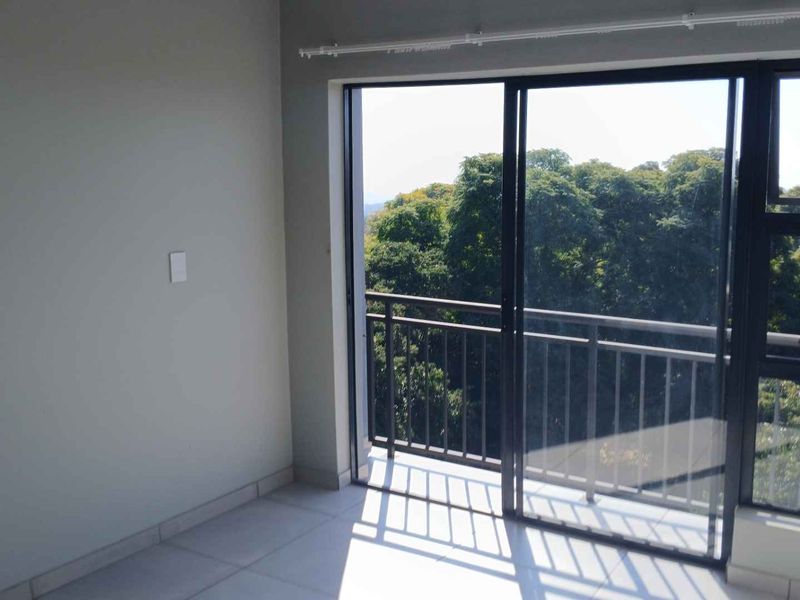 An empty bedroom featuring tiled flooring and a sliding glass door that opens onto a balcony with a scenic tree-lined view.
