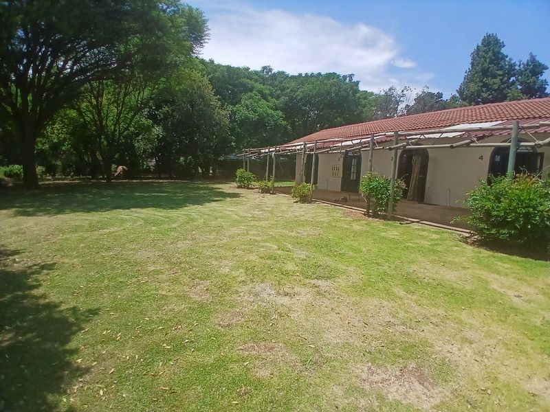 A rear view of the cottage featuring a paved patio area covered by a timber pergola, overlooking a spacious garden with mature trees.