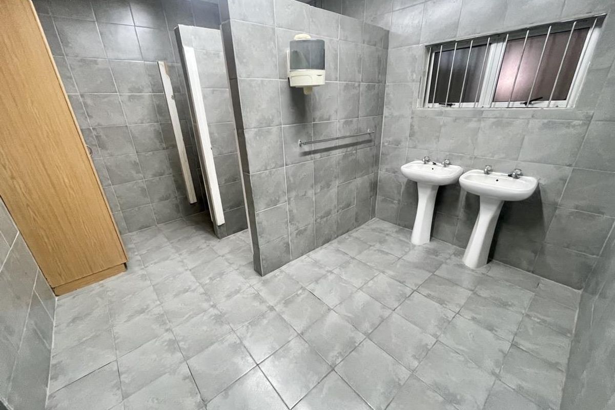 A functional office restroom featuring dual pedestal sinks, tiled walls and flooring, and partitioned cubicle areas.