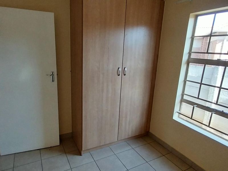 A clean, empty bedroom featuring neutral tiled flooring, a built-in wooden wardrobe, and a window fitted with security bars.