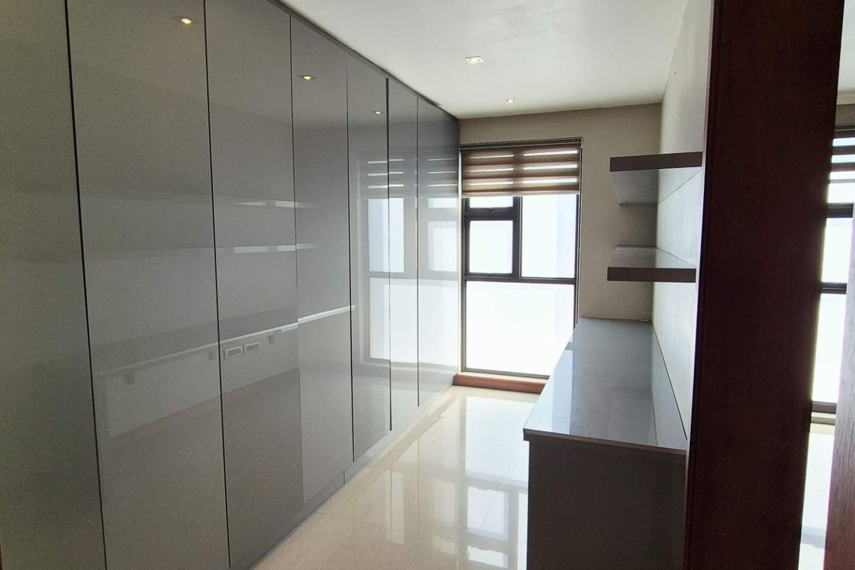 A modern, empty bedroom featuring extensive high-gloss built-in cupboards, a built-in desk, tiled flooring, and recessed lighting.
