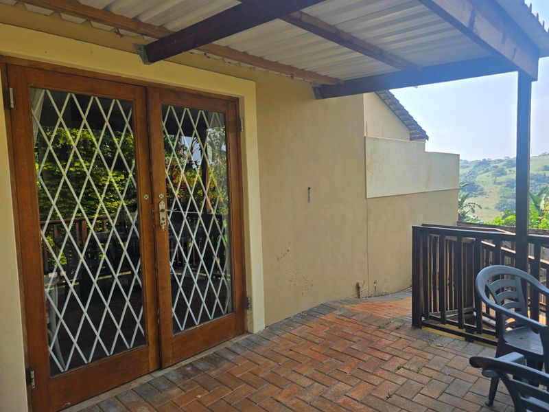 A covered patio area featuring brick paving, timber-framed glass double doors with security trellis, and a timber balustrade overlooking the surrounding landscape.