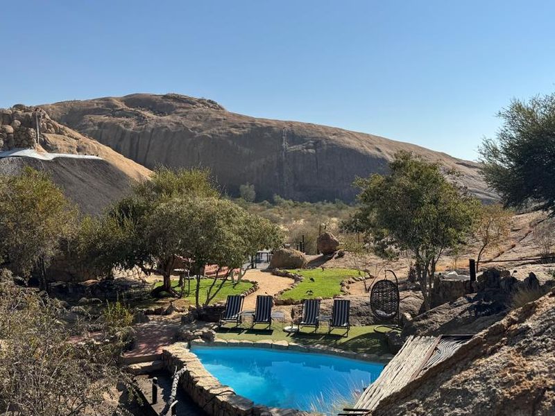 A scenic outdoor pool area at a lodge, featuring natural rock landscaping, a thatched roof structure, and expansive mountain views.