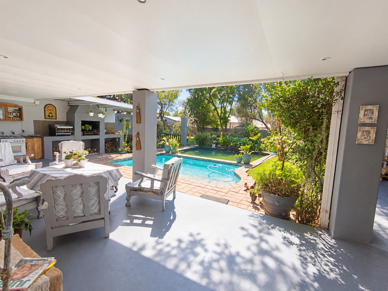 Covered outdoor patio featuring a built-in braai area overlooking a swimming pool with paved surround and established garden.