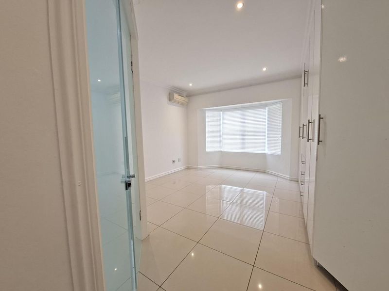 A bright, empty bedroom featuring large tiled floors, extensive built-in cupboards, a bay window with blinds, and a wall-mounted air conditioning unit.