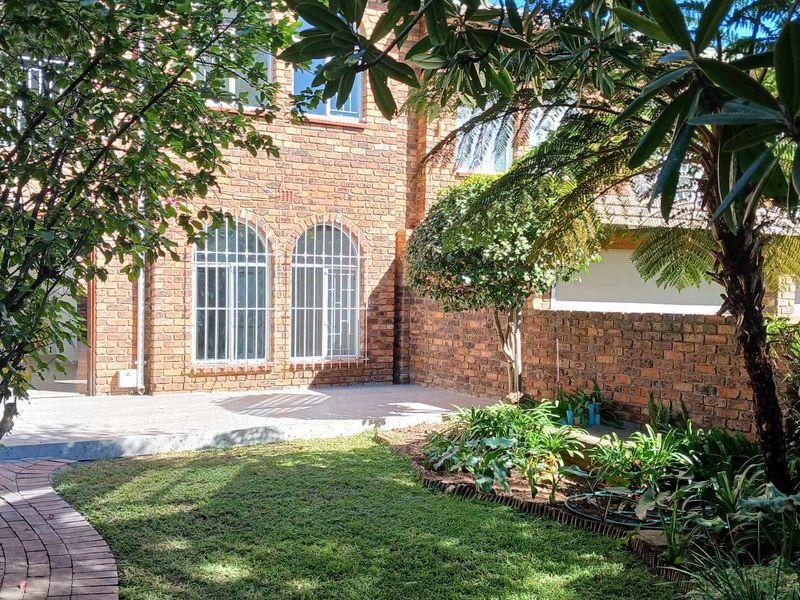 A private rear garden featuring a face brick townhouse facade, arched windows with security bars, a paved patio area, and established landscaping.