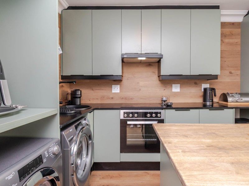 Modern kitchen featuring sage green cabinetry, black stone countertops, wood-look splashback, and integrated appliances including an oven and hob.