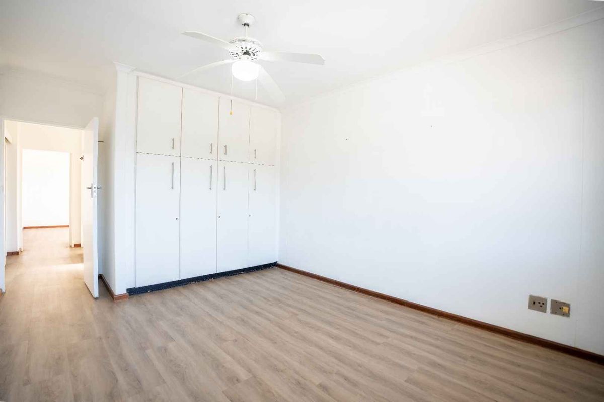 A bright, empty bedroom featuring light-toned laminate flooring, built-in white cupboards, and a ceiling fan, offering a clean canvas for personalization.