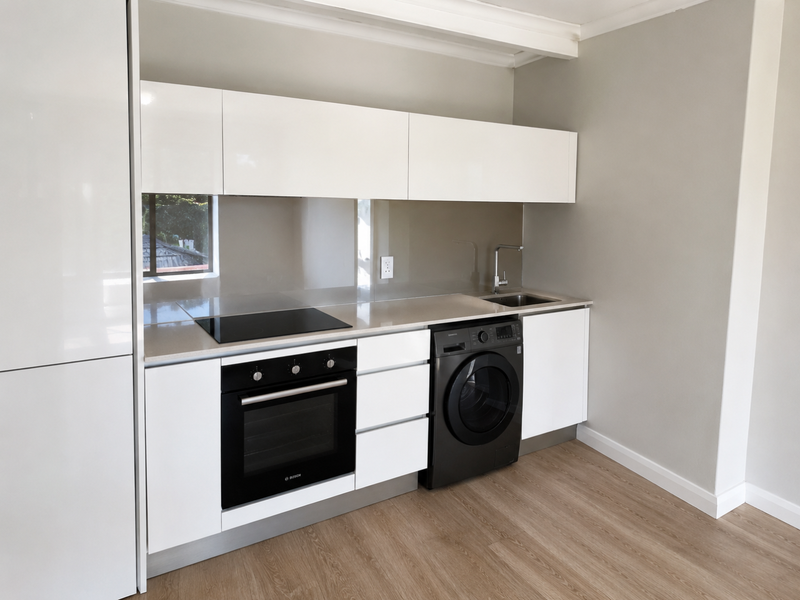 Modern, minimalist kitchen featuring sleek white cabinetry, stone countertops, integrated electric hob, oven, and a clean glass splashback in a neutral space.