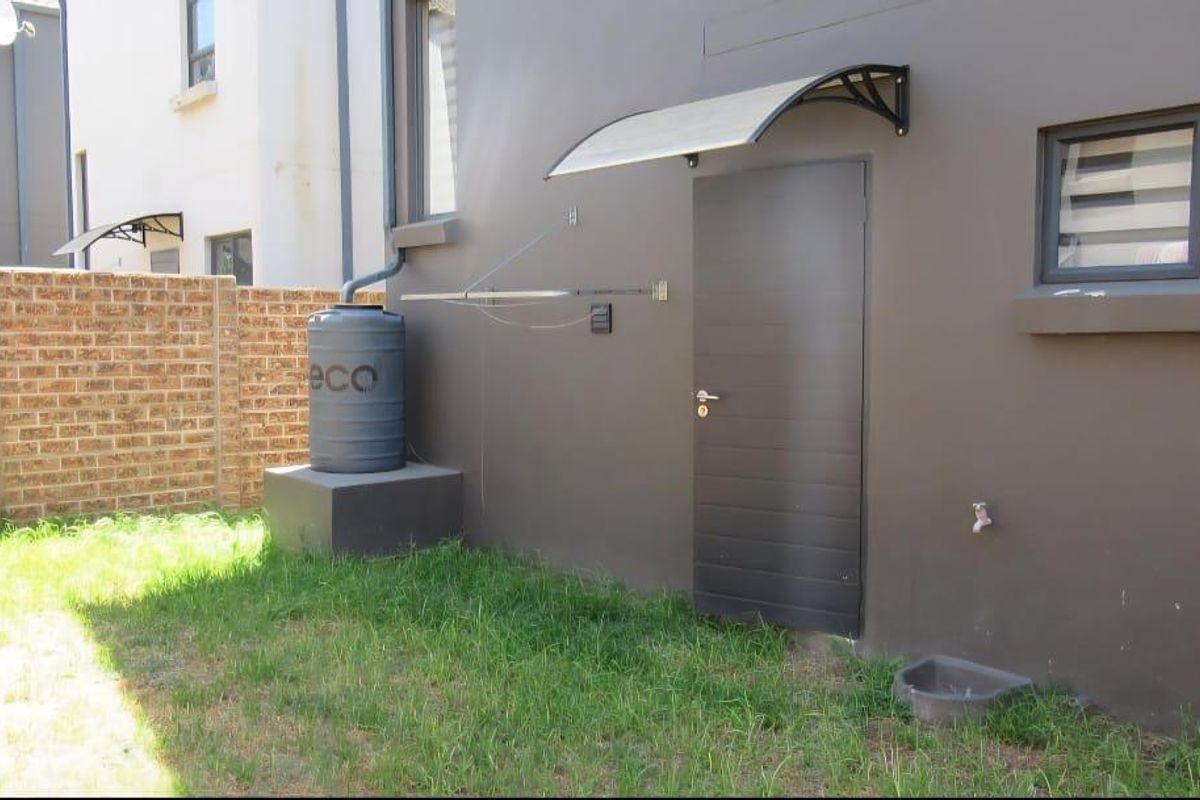 Rear exterior view of the townhouse featuring a small garden, water tank, wall-mounted drying rack, and a side access door with a protective awning.