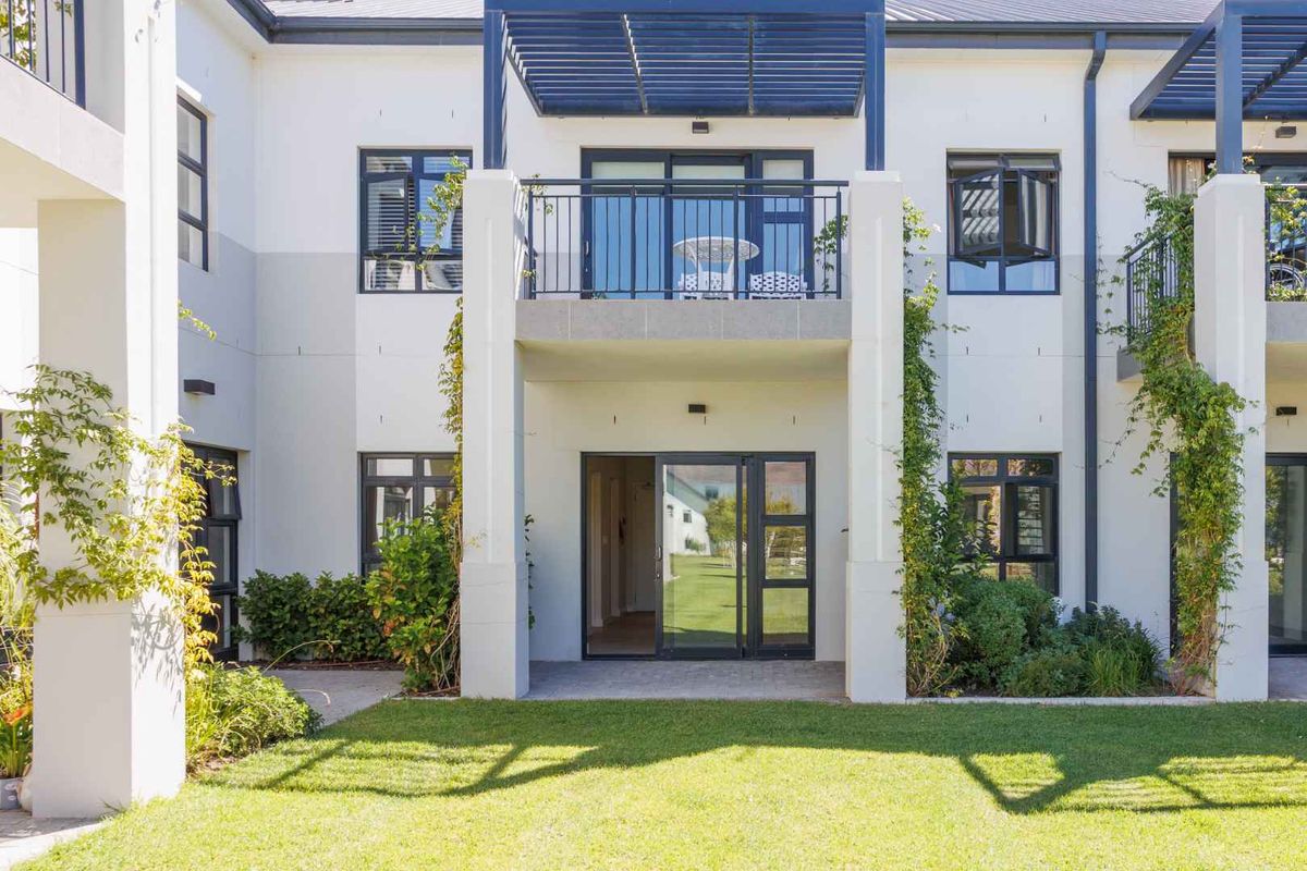Rear view of the apartment building featuring a ground floor patio, upper balcony with pergola, and well-maintained garden area.