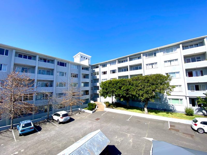 An elevated view of the apartment complex exterior, featuring a paved parking area, landscaped garden spaces, and multi-story residential building facade.