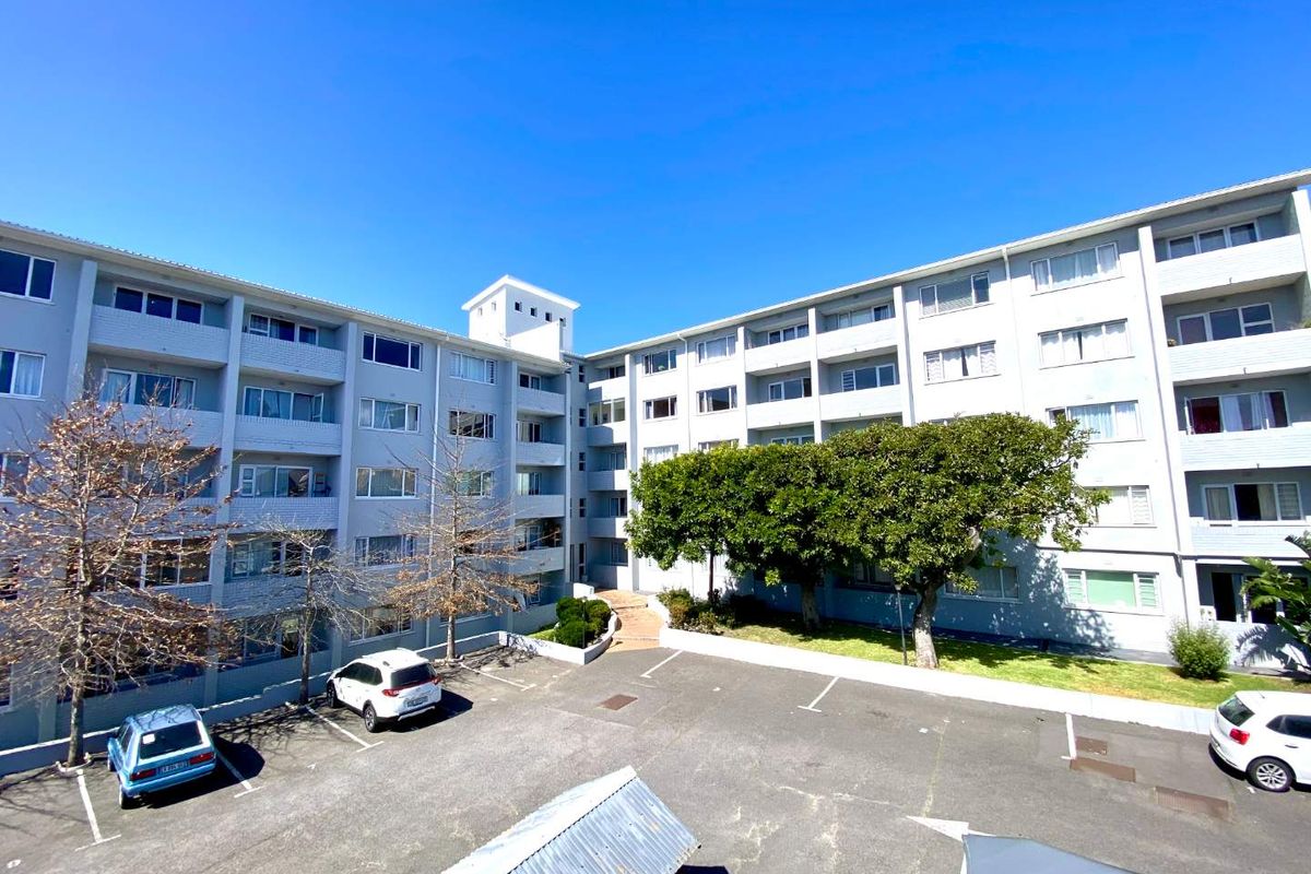 An elevated view of the apartment complex exterior, featuring a paved parking area, landscaped garden spaces, and multi-story residential building facade.