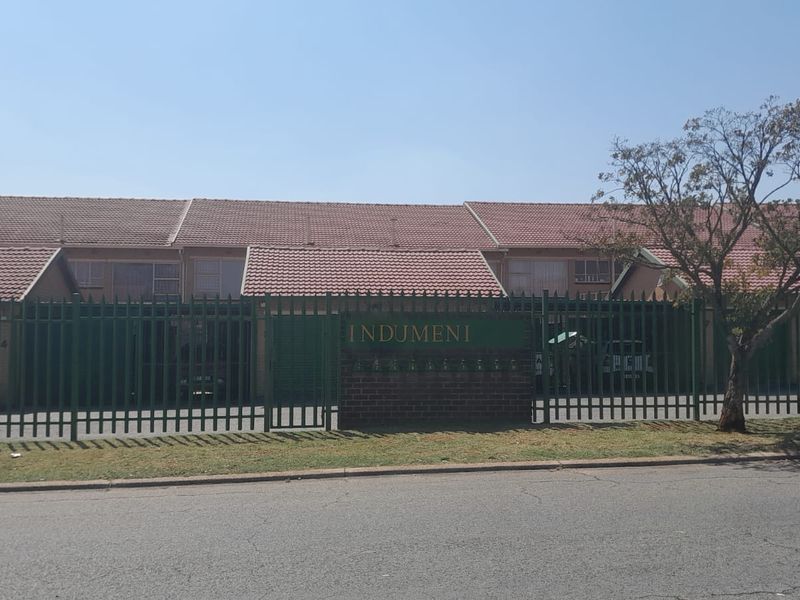 Front exterior view of the Indumeni townhouse complex featuring a tiled roof, perimeter security fencing, and a brick entrance signage wall.