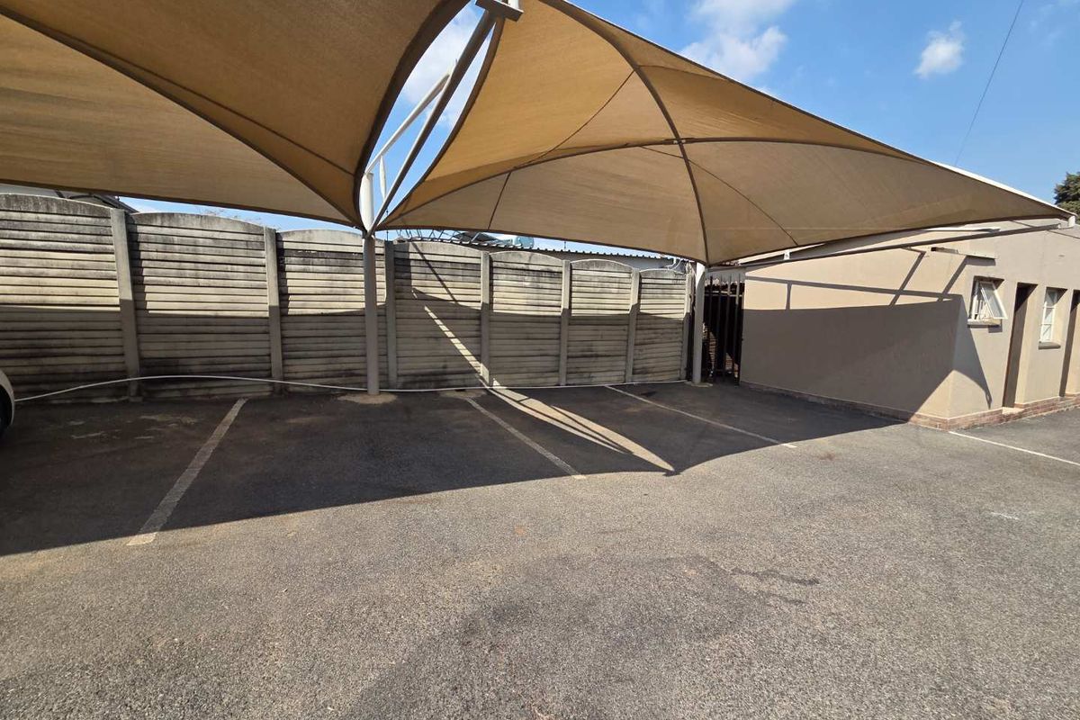 Outdoor parking area featuring a large shade sail structure, asphalt paving, and a secure precast concrete boundary wall.