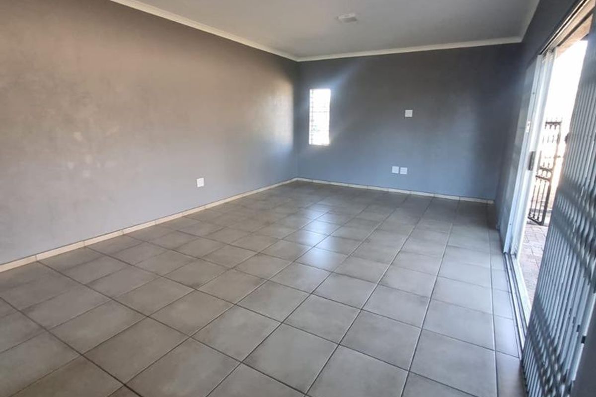 An empty, open-plan living area featuring neutral grey walls, ceramic floor tiles, and a sliding glass door with a security gate leading outdoors.