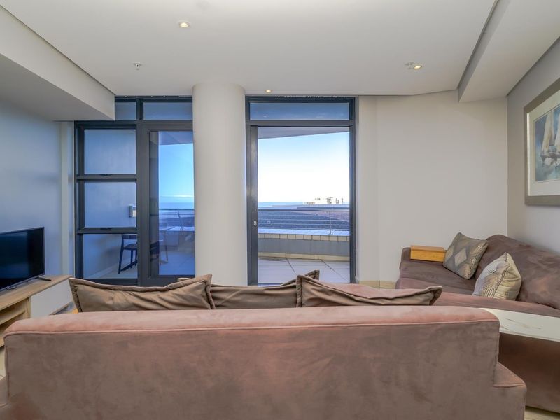 A bright living room featuring tiled flooring, recessed ceiling lights, and large sliding glass doors that provide access to a balcony with ocean views.