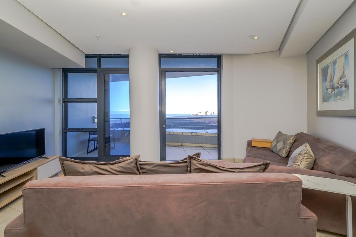 A bright living room featuring tiled flooring, recessed ceiling lights, and large sliding glass doors that provide access to a balcony with ocean views.