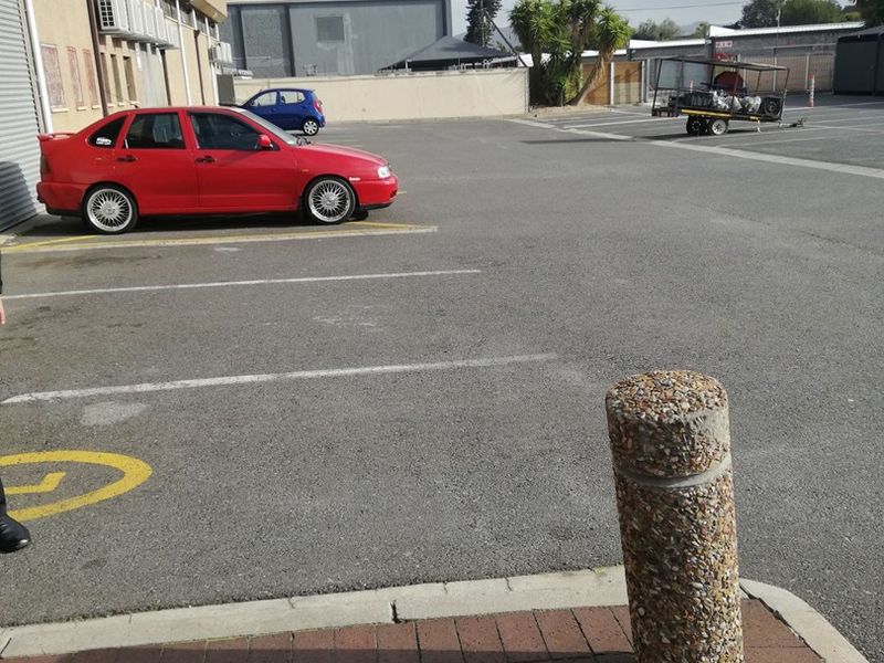 Exterior view of the office property featuring an asphalt parking area, concrete bollards, and brick paving with external air conditioning units.